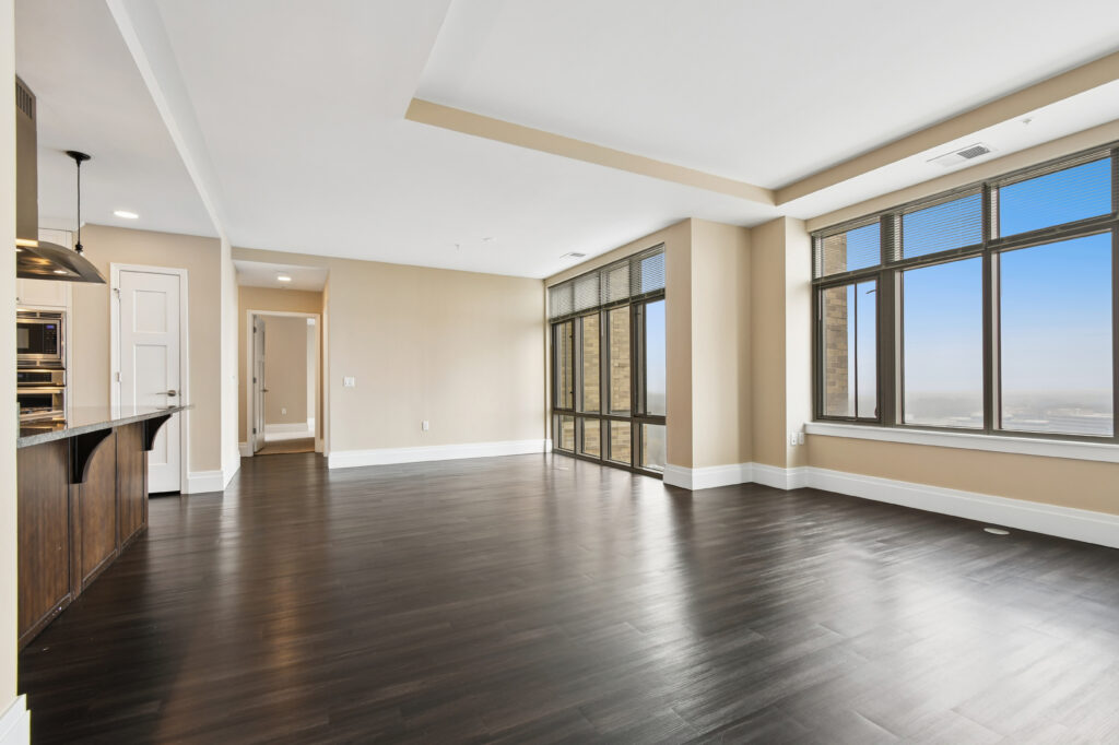 Spacious, modern apartment with dark wood floors, large windows letting in natural light, beige walls, and an open kitchen with stainless steel appliances and pendant lighting.