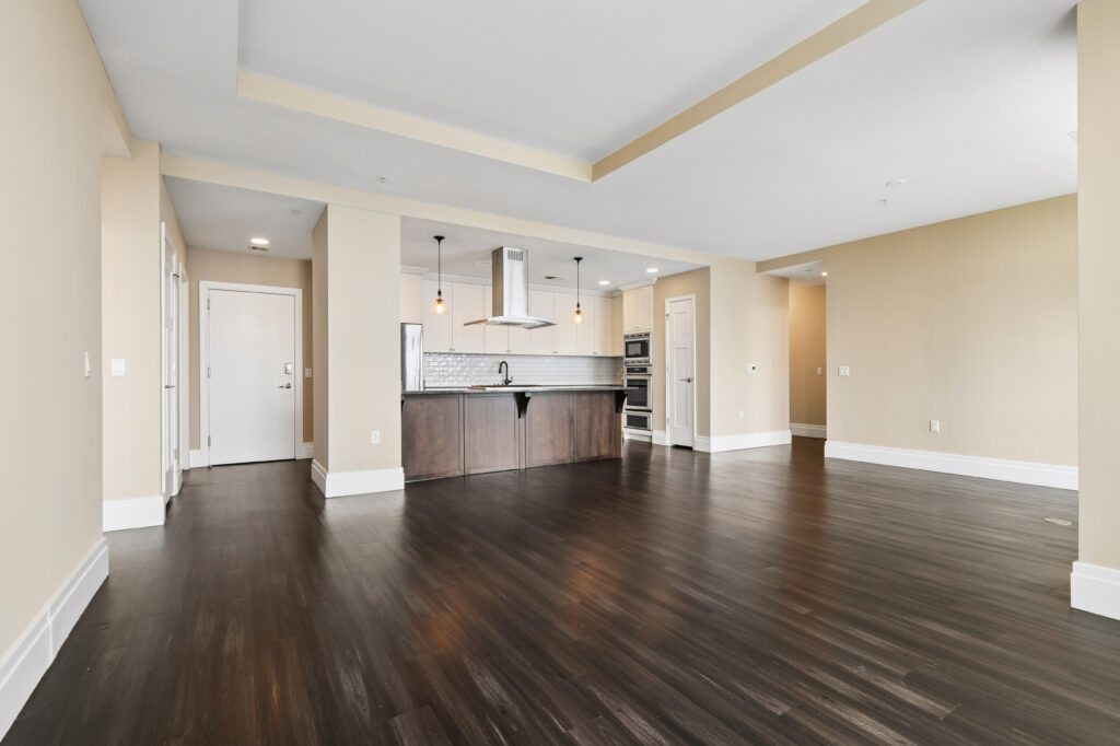 Spacious modern apartment with dark hardwood floors, beige walls, recessed ceiling, and an open-concept kitchen featuring a large island, pendant lights, white cabinetry, and stainless steel appliances.