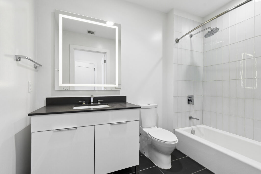 Modern bathroom with white walls and fixtures, a rectangular sink with black countertop, a large illuminated mirror above, a toilet, and a bathtub with a glass shower curtain rod and white tiled wall.