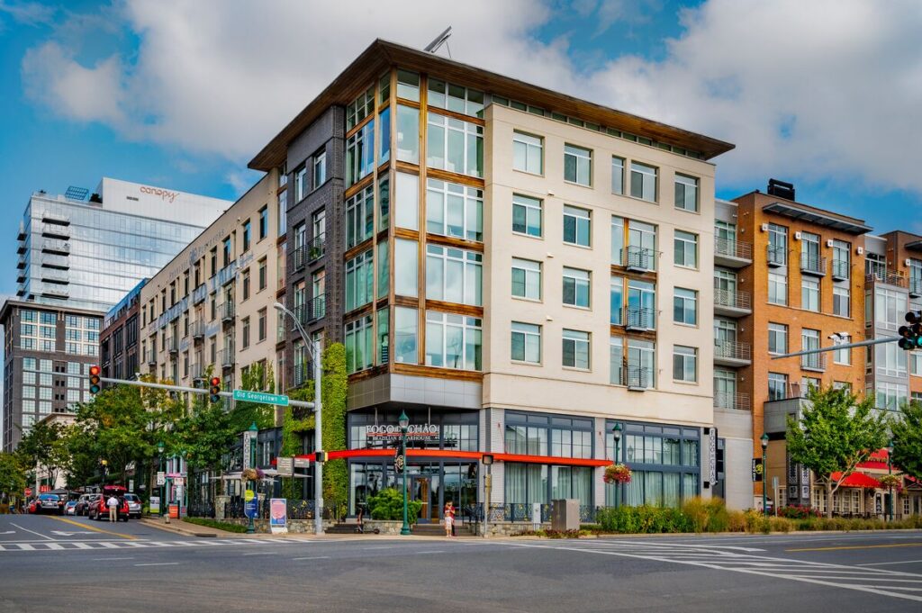 A modern, multi-story apartment building with retail stores on the ground level sits on a city street corner, surrounded by traffic lights, trees, and other contemporary buildings under a partly cloudy sky.