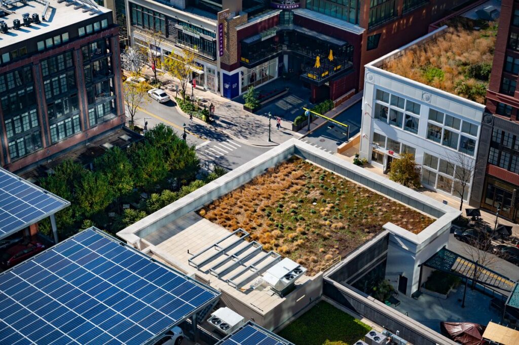 Aerial view of a city block showing a building with a green roof covered in grass and plants, surrounded by solar panels, trees, streets, and pedestrians in an urban environment.