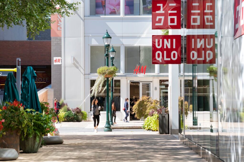 Outdoor mall scene with people walking past shop entrances for H&M and UNIQLO, surrounded by plants, street lamps, and red store signs on a sunny day.
