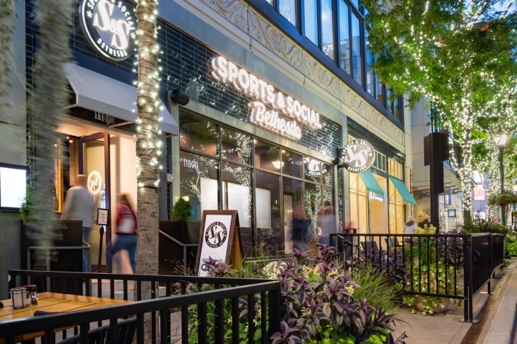 Outdoor view of a lively restaurant and bar with Sports & Social Bethesda signage, outdoor seating, decorative plants, and string lights wrapped around trees, as people walk by on the sidewalk.