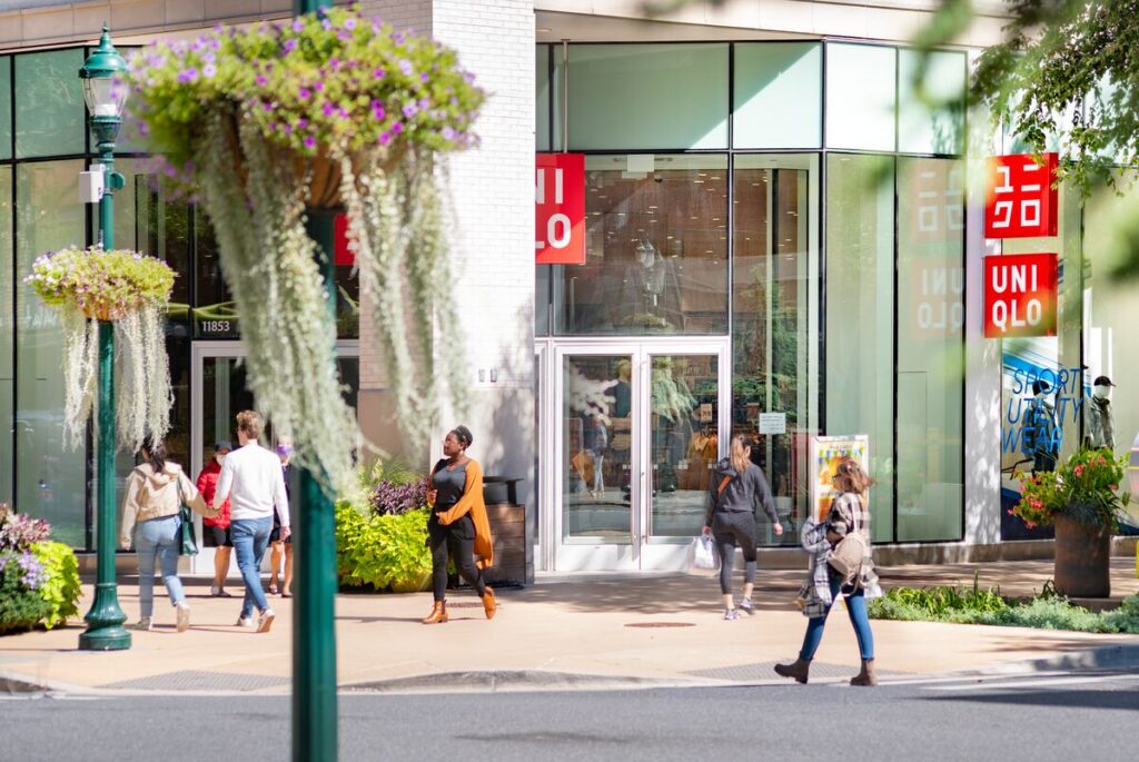 People walk past a UNIQLO store with large glass windows on a sunny day, surrounded by greenery and hanging flower baskets. Some are entering or exiting the store, while others walk along the sidewalk.