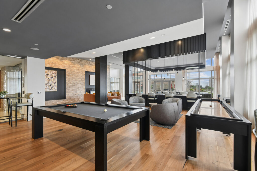 Modern lounge with a pool table and shuffleboard, wooden floors, gray seating area, large windows letting in natural light, and contemporary decor with a stone accent wall.