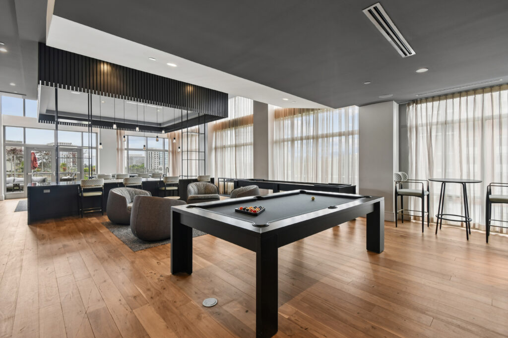 Modern lounge area with a pool table in the foreground, comfortable seating, tall windows with sheer curtains, and high tables with stools along the side, all on a wood floor with neutral, contemporary decor.