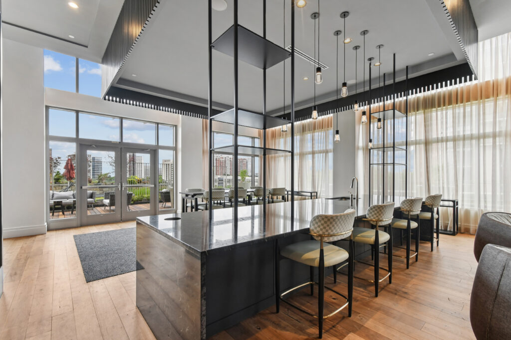 Modern communal kitchen and dining area with a large black island, stylish pendant lights, cushioned barstools, floor-to-ceiling windows, light wood floors, and city buildings visible outside.