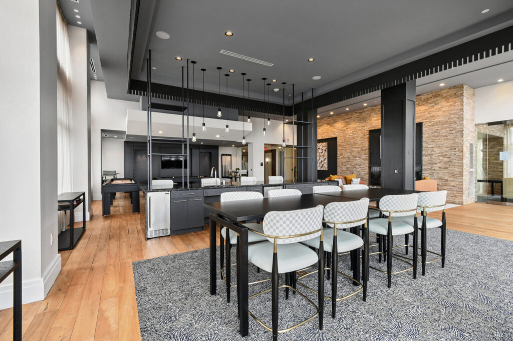 Modern communal dining and lounge area with a black dining table, eight white cushioned chairs, stylish pendant lights, a gray rug, hardwood floors, and a mix of black and stone accent walls.