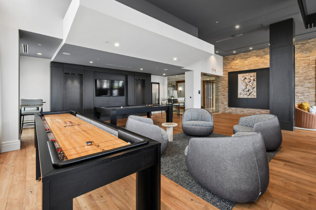 Modern lounge area with a shuffleboard table, pool table, round gray chairs, a TV mounted on a dark wall, wood flooring, and contemporary decor, including a stone accent wall with framed artwork.