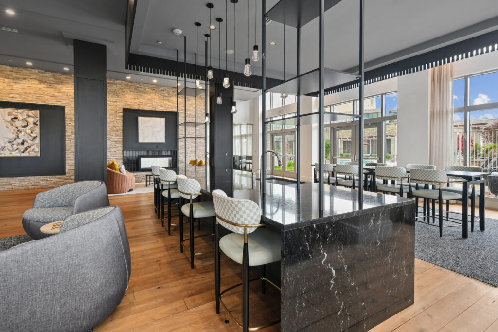 Modern communal space with a marble island, upholstered chairs, and pendant lights. Large windows let in natural light, with cozy seating, wood floors, and stone accent walls creating a stylish, inviting atmosphere.