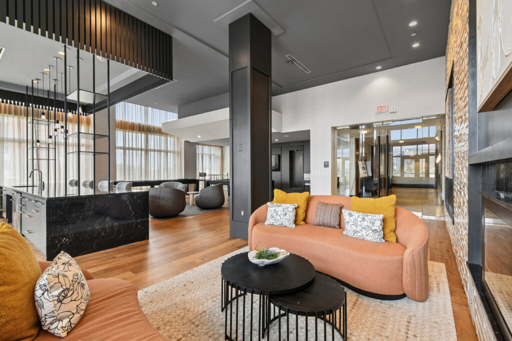 Modern lounge area with peach sofas, yellow and patterned pillows, round black coffee tables, hardwood floors, and large windows letting in natural light. A black marble counter and contemporary decor are visible in the background.