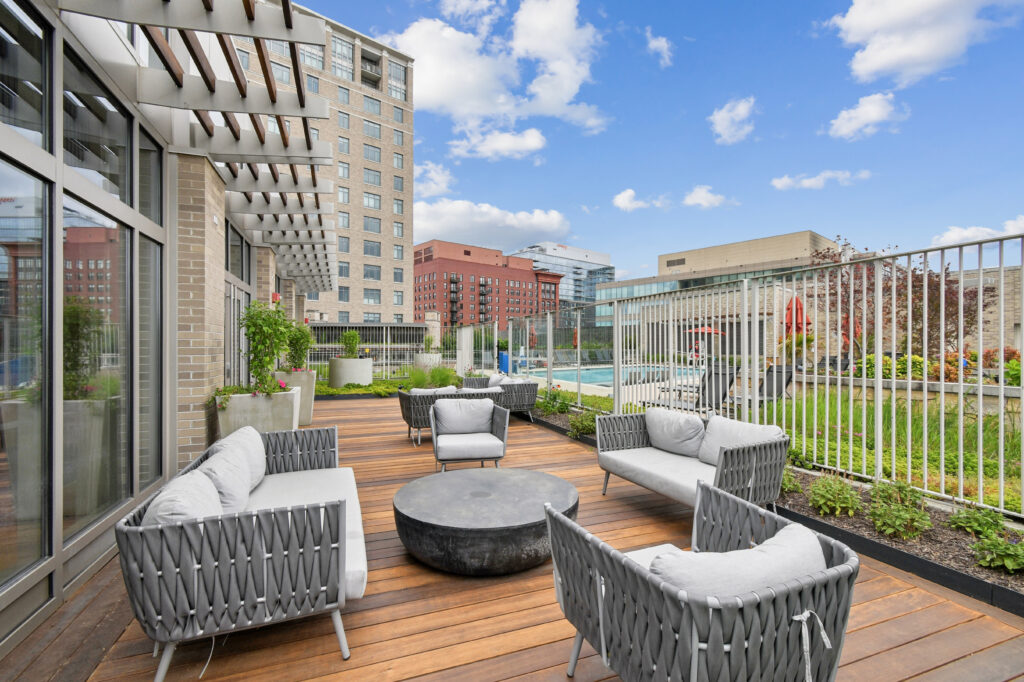 Modern rooftop patio with woven gray seating, round black coffee table, wooden deck, and green landscaping; high-rise buildings and blue sky with clouds in the background.