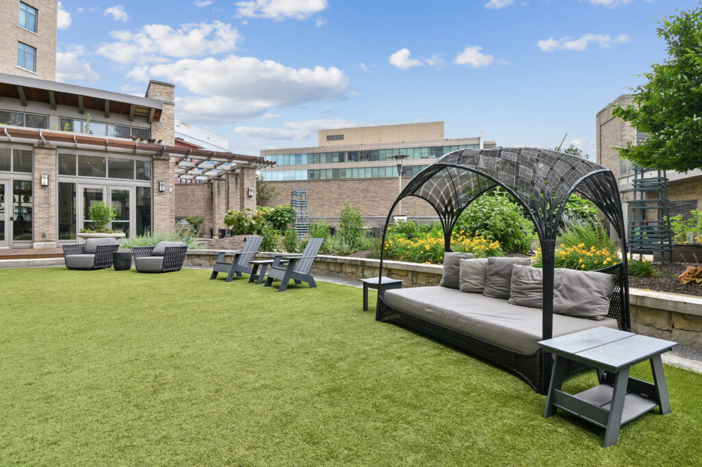 An urban rooftop patio with artificial turf, outdoor seating including a cabana bed and lounge chairs, and lush landscaping.