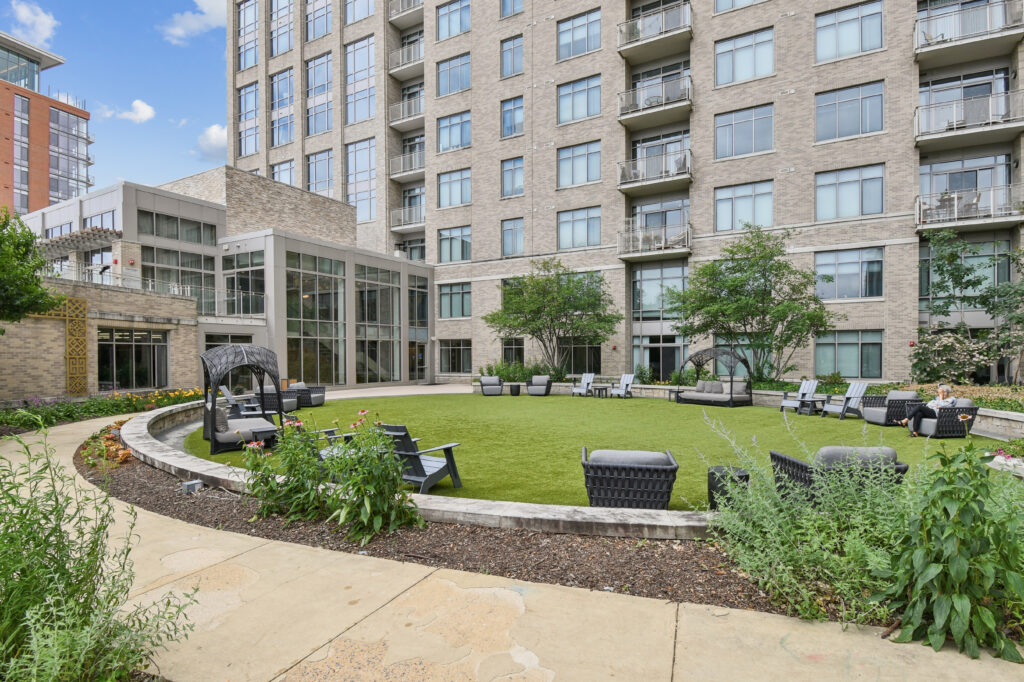 A modern apartment building with balconies overlooks a landscaped courtyard featuring artificial grass, seating areas, outdoor furniture, and flowering plants on a sunny day.