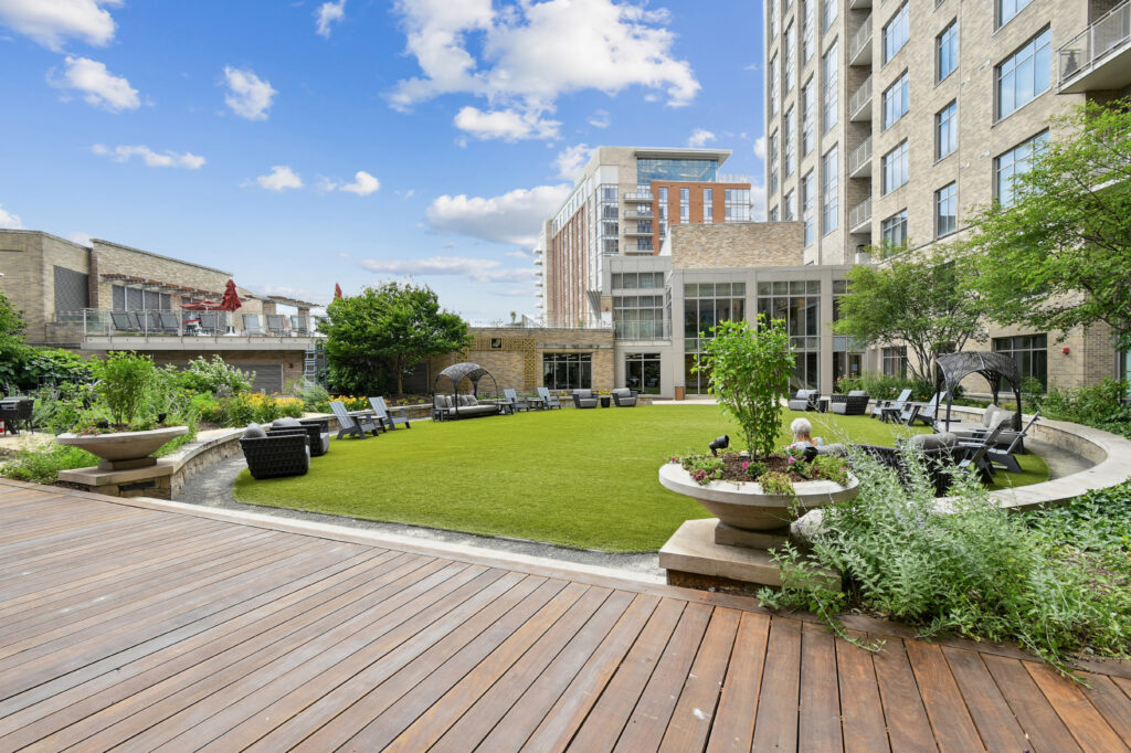 A landscaped outdoor courtyard with artificial grass, lounge chairs, wicker furniture, planters, and wooden decking, surrounded by modern apartment buildings under a blue sky with scattered clouds.