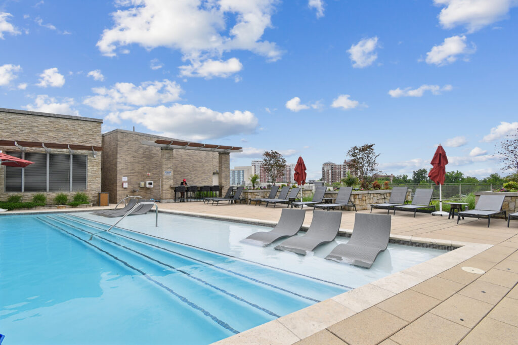 A modern outdoor swimming pool with submerged lounge chairs, surrounded by a sun deck with red umbrellas and lounge chairs, overlooking a cityscape under a partly cloudy sky.