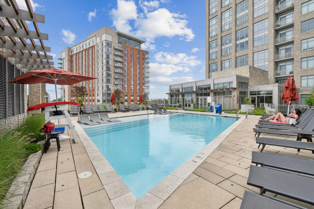 Outdoor swimming pool at an apartment complex, surrounded by lounge chairs, umbrellas, and tall modern buildings under a blue sky with scattered clouds. A person is relaxing on a chair by the pool.