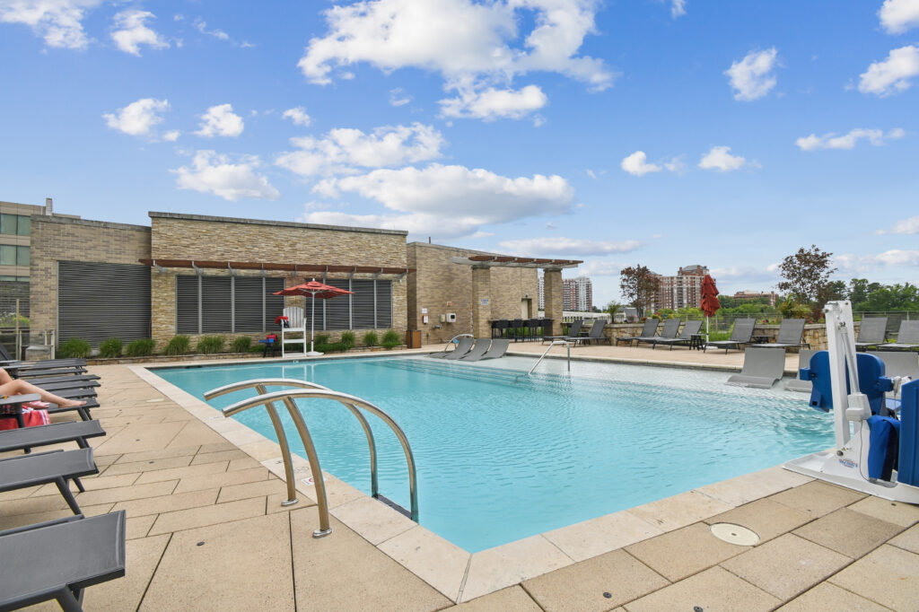 Outdoor swimming pool with clear blue water, surrounded by lounge chairs, a few tables with red umbrellas, and a modern building in the background under a partly cloudy sky.