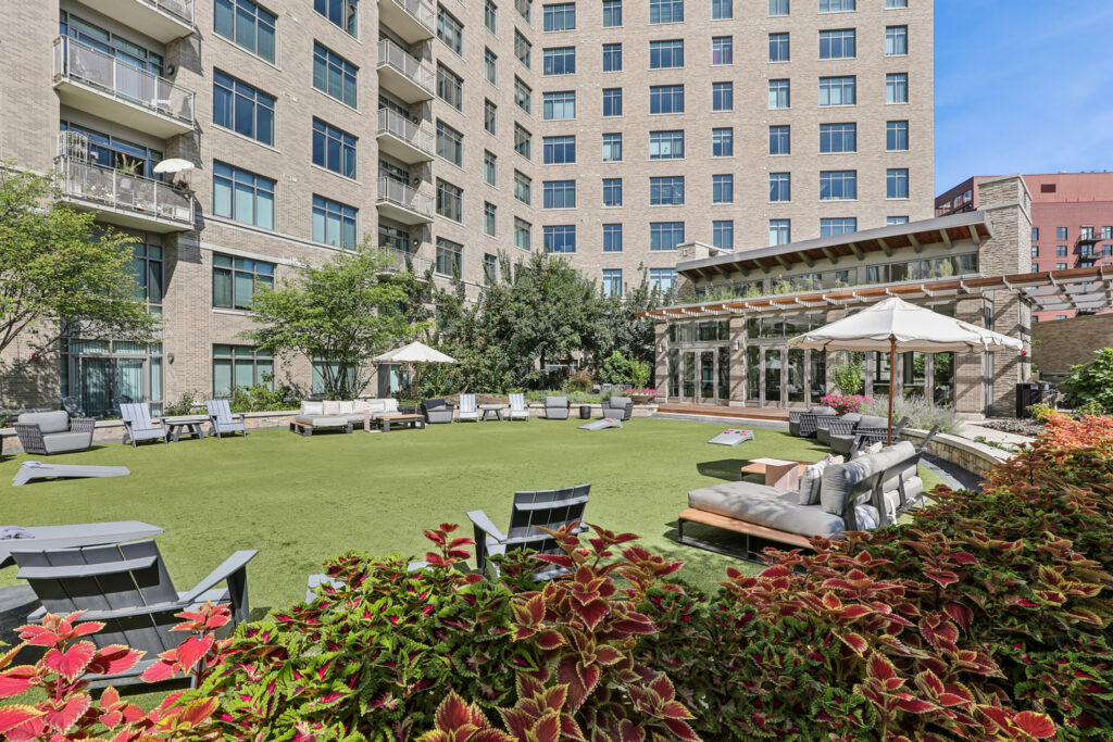 Outdoor courtyard of an apartment building with green lawn, lounge chairs, umbrellas, cornhole boards, and lush plants. Tall buildings and a glass-enclosed patio are in the background.