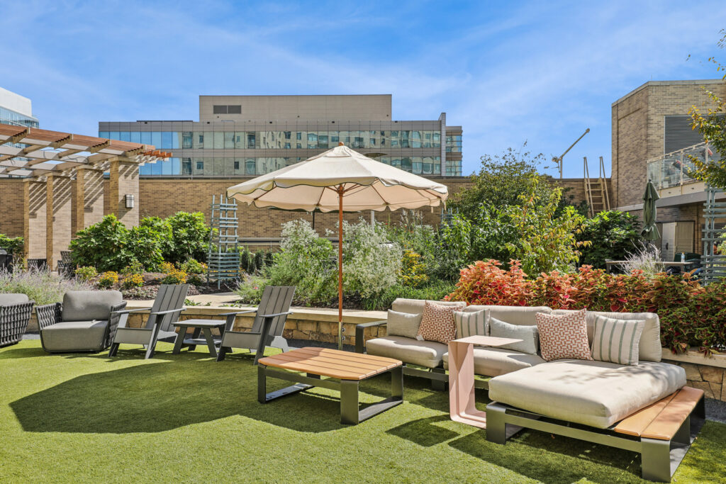 Outdoor rooftop lounge with cushioned sofas, wooden chairs, a table, and a large umbrella, surrounded by greenery and modern buildings under a blue sky.
