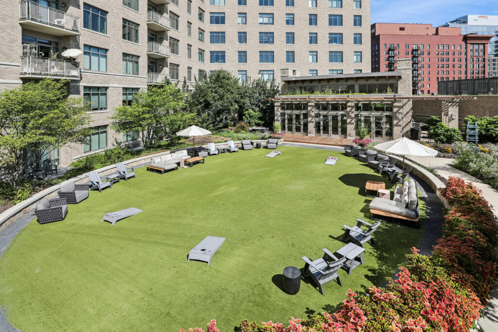 A rooftop lawn with scattered patio furniture, bean bag toss boards, shade umbrellas, and a view of surrounding city buildings. Shrubs and flowers border the green space.