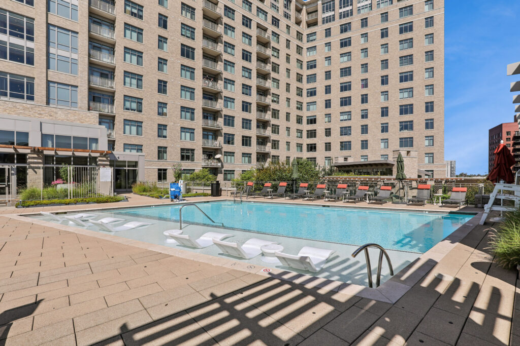 Outdoor pool area at an apartment complex, featuring in-water lounge chairs, orange-cushioned chairs along the deck, and a tall tan building in the background on a clear sunny day.