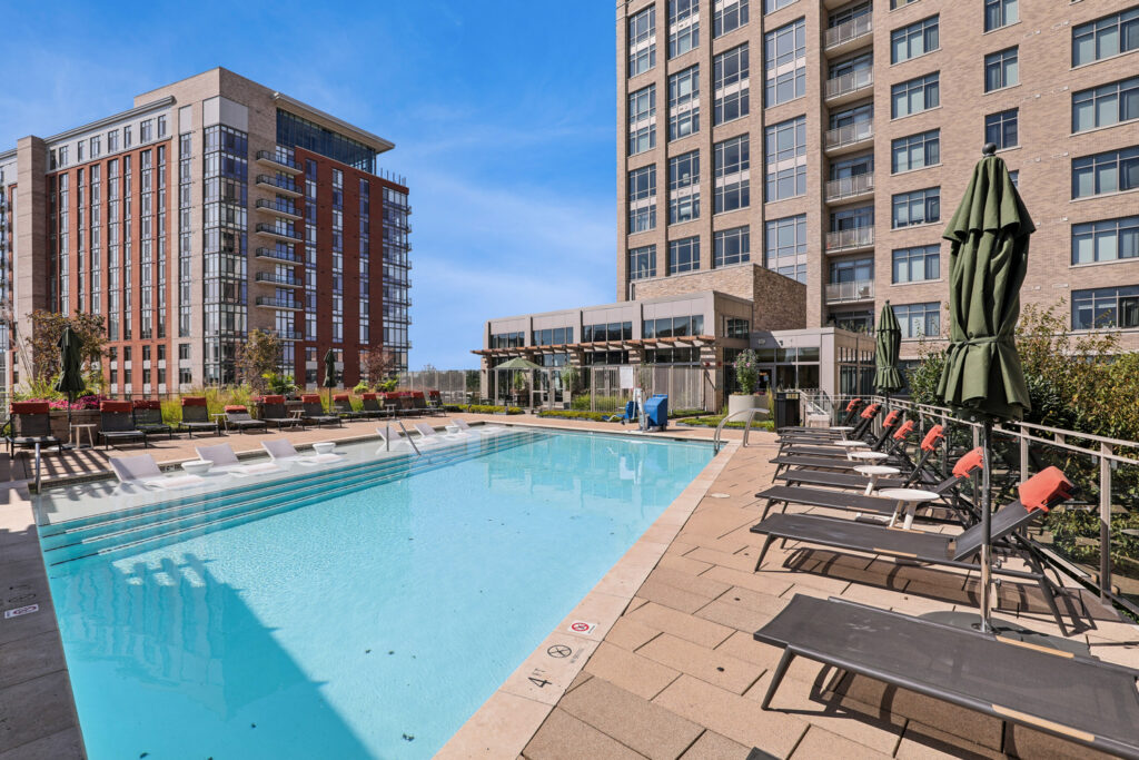 Modern apartment building outdoor pool area with lounge chairs, umbrellas, and potted plants. Tall buildings surround the pool, and the sky is clear and blue.
