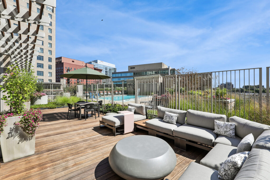 Outdoor rooftop lounge with cushioned seating, potted plants, a table with chairs and an umbrella, wooden deck, and a fenced swimming pool in an urban setting under a clear blue sky.